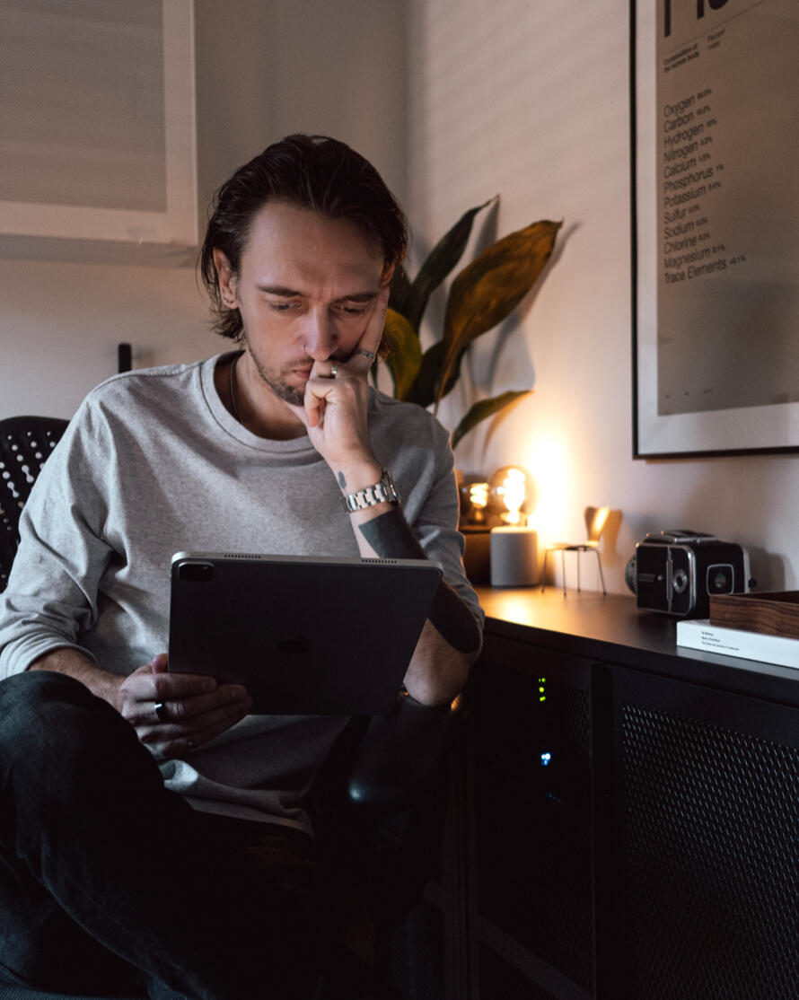 A photograph of Ben sitting in an urban area and looking off-camera. He's wearing winter clothes: a beanie hat, sunglasses, a woolly jumper and a technical coat.