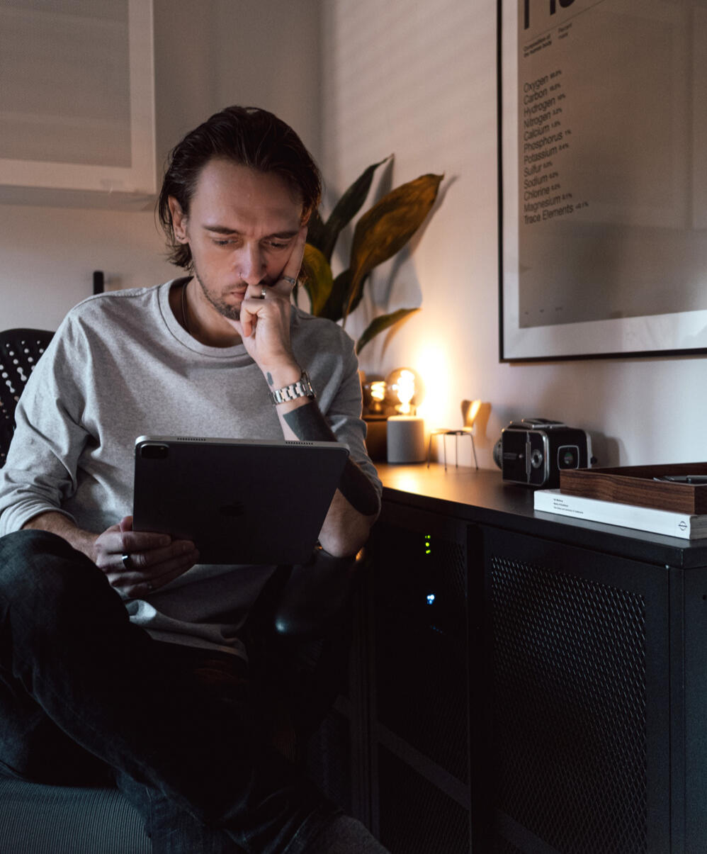 A photograph of a minimal wooden desk in a bright room, featuring a wall-mounted monitor, a tall, potted cactus, and a large, white desktop speaker.
