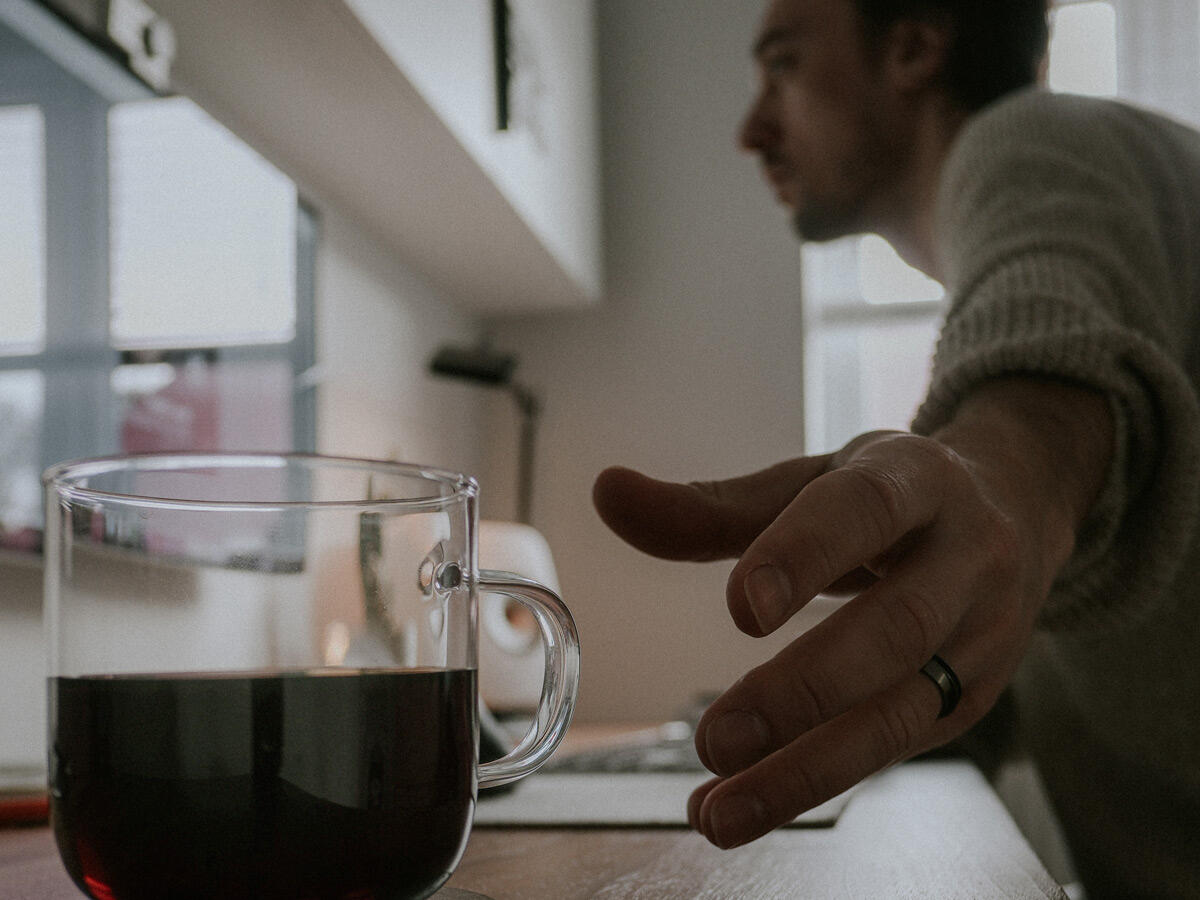 A photograph of Ben sitting in an urban area and looking off-camera. He's wearing winter clothes: a beanie hat, sunglasses, a woolly jumper and a technical coat.