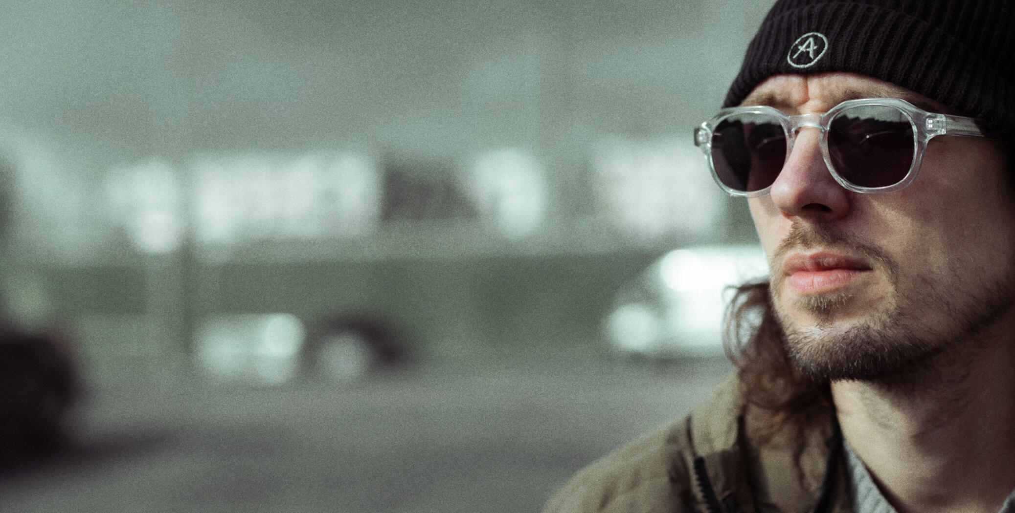 A photograph of Ben sitting in an urban area and looking off-camera. He's wearing winter clothes: a beanie hat, sunglasses, a woolly jumper and a technical coat.