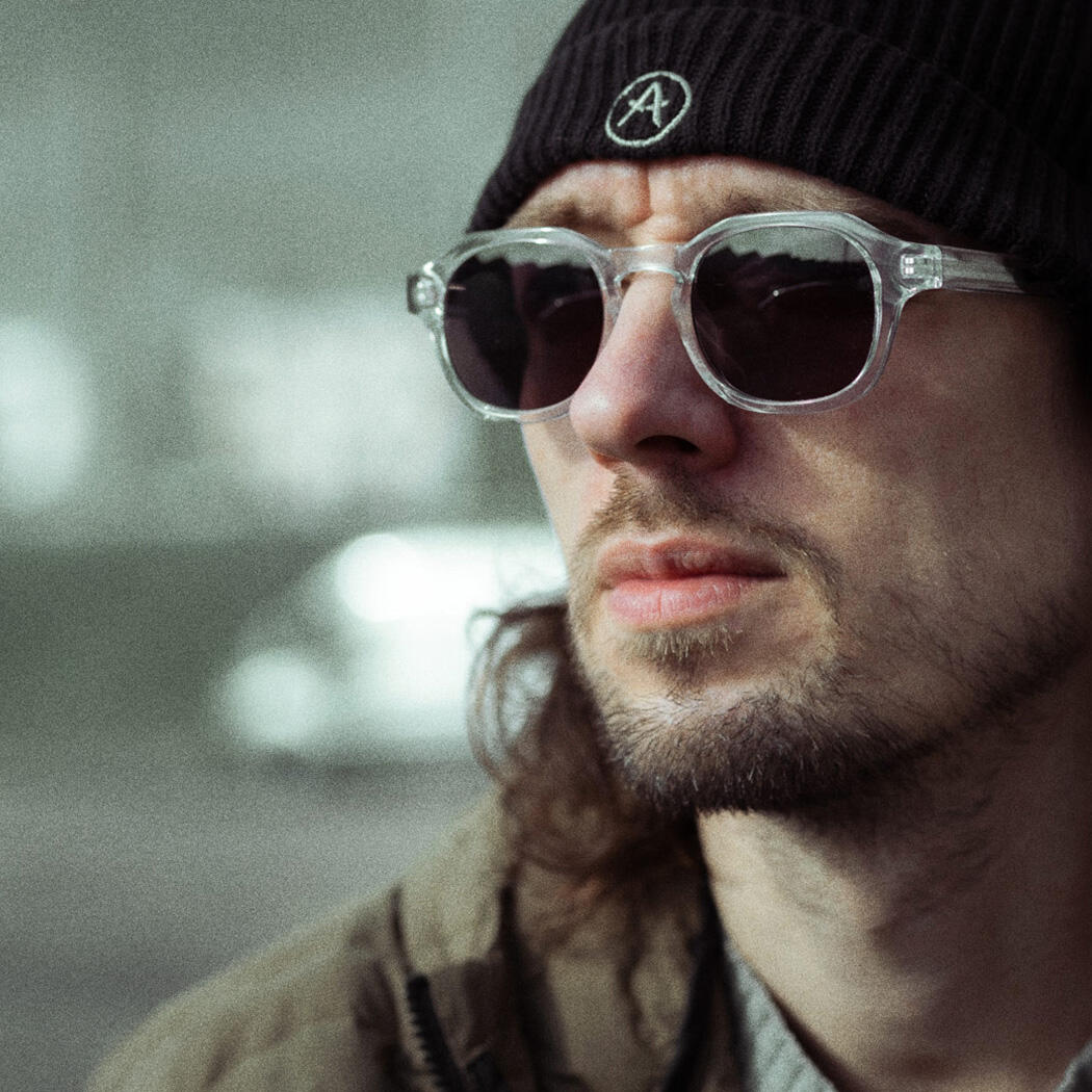 A photograph of Ben sitting in an urban area and looking off-camera. He's wearing winter clothes: a beanie hat, sunglasses, a woolly jumper and a technical coat.